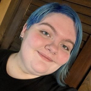 A photo of a person taken at a slight angle. They are white and have short, medium blue hair and blue eyes. They are smiling.